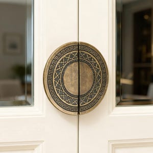 Puede incluir: Un par de puertas blancas con un tirador circular de color bronce. El tirador presenta un intrincado patrón geométrico alrededor del borde y el centro, añadiendo un toque de elegancia a las puertas.