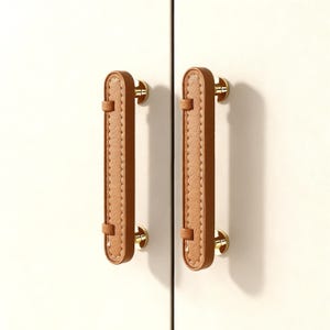 May include: Two cabinet pulls with a light brown leather-effect centre and gold-coloured hardware. The pulls are vertically oriented and attached to a white cabinet door. The centre section has a stitched design.