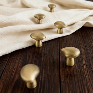 May include: Several mushroom-shaped cabinet knobs in a warm brass finish. The knobs are displayed on a dark wooden surface with a cream-coloured fabric. Each knob has a smooth, rounded top and a cylindrical base. The knobs are made of brass.