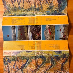 May include: Folded vintage map featuring a detailed illustration of a mountain range with peaks and valleys. The map includes aerial tramways and text labels in multiple languages. The color palette includes shades of blue, green, yellow, and brown.