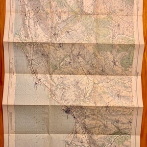 May include: Folded vintage map with detailed topographical features, including coastlines, roads, and buildings. The map is printed on aged paper, showing signs of wear. The map is in shades of green, brown, and blue, with text in the upper right corner.