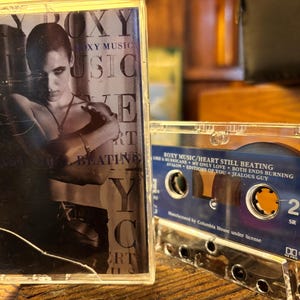 Puede incluir: Una cinta de cassette vintage del álbum "Heart Still Beating" de Roxy Music. La carcasa transparente muestra el título del álbum y la lista de canciones. La portada presenta una imagen en blanco y negro. La cinta está sobre una superficie de madera.