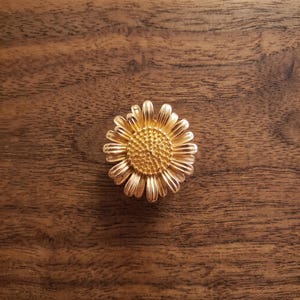 May include: A gold-toned metal brooch shaped like a daisy flower. The piece features a textured, raised central disc surrounded by numerous slender, radiating petals with vertical grooves, creating a detailed floral appearance.