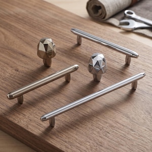 May include: A collection of silver-toned cabinet hardware including two cylindrical bar pulls and two faceted, geometric knobs. The hardware features a polished metallic finish with smooth, rounded ends on the pulls and multi-faceted surfaces on the knobs.