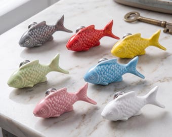 Può includere: Una collezione di sette pomelli per mobili in ceramica a forma di pesce in vari colori, tra cui grigio, rosso, giallo, verde chiaro, azzurro, rosa e bianco. Ogni pomello presenta una texture a scaglie dettagliata e una base in metallo.