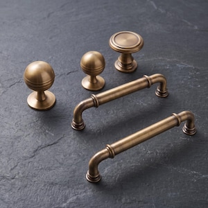 May include: A collection of antique brass hardware including two cylindrical cabinet pulls with curved ends and three distinct cabinet knobs. The knobs feature varied shapes, including spherical and flat-topped designs, all finished in a brushed metallic tone.
