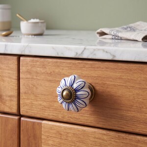 Pu&ograve; includere: Un pomello rotondo in ceramica per mobili con base bianca, motivi a petali dipinti in blu e un accento centrale in metallo color ottone antico. Il pomello &egrave; montato su un frontale di cassetto in legno chiaro con venature visibili.