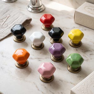 Può includere: Una collezione di nove pomelli esagonali per mobili in vari colori lucidi, tra cui rosso, nero, bianco, giallo, arancione, viola, rosa e verde. Ogni pomello presenta una superficie superiore liscia e sfaccettata su una base in ottone anticato.
