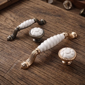 May include: Two arched drawer pulls and two round knobs featuring white ceramic centers with ribbed textures. The hardware components have metallic bases with a dark bronze finish, showcasing fluted detailing on the ends and edges.
