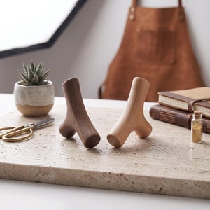 May include: Two Y-shaped wooden massage tools, one crafted from dark walnut wood and the other from light maple wood, standing upright on a textured stone surface. The tools feature smooth, rounded ends and a ergonomic design for grip.