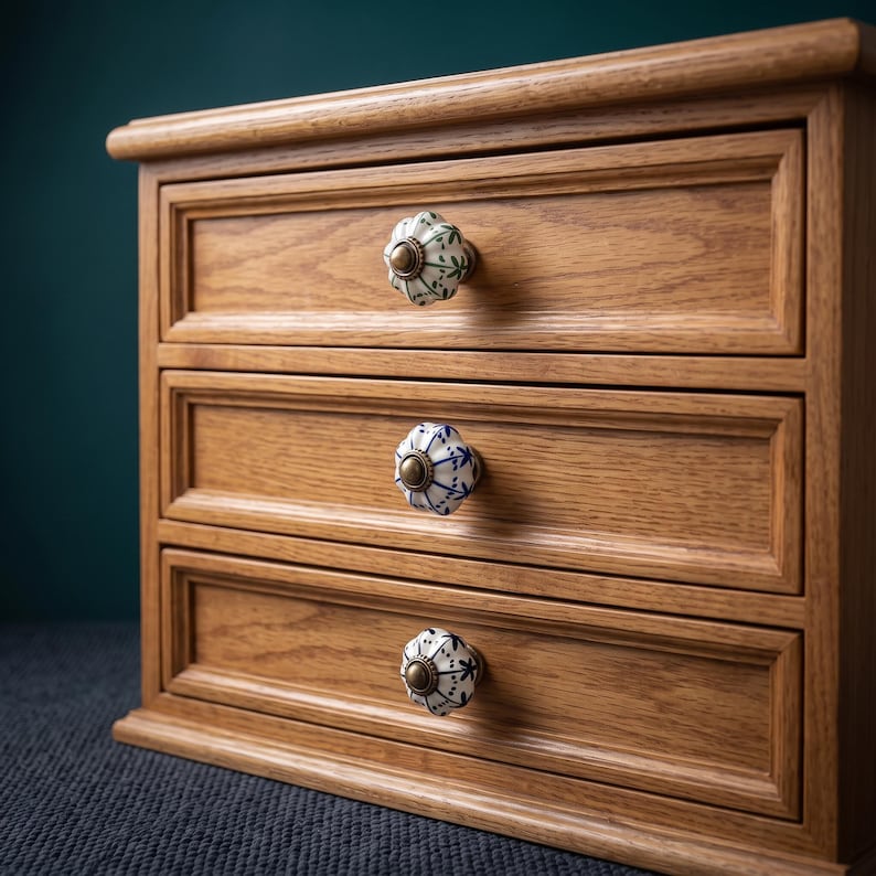 Pu&ograve; includere: Una cassettiera in legno a tre cassetti, ognuno dotato di un pomello in ceramica distinto. Il pomello superiore presenta motivi verdi, quello centrale motivi blu e quello inferiore motivi neri. Ogni pomello &egrave; rotondo, scanalato e ha un centro in ottone.