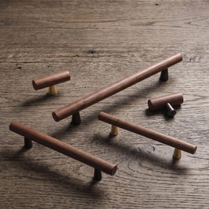 May include: A collection of five cabinet pulls and knobs featuring cylindrical dark brown wood handles. The hardware includes three long horizontal pulls and two smaller T-shaped knobs, each mounted on small conical bases in brass or dark metal.