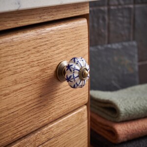 Pu&ograve; includere: Un pomello per mobili in ceramica rotondo e scanalato, caratterizzato da una base bianca con un motivo floreale blu. Il pomello &egrave; fissato su un frontale di cassetto in legno chiaro e include una base in metallo color ottone e una vite centrale.