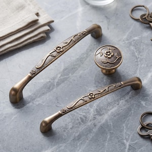 Può includere: Due maniglie curve per armadietto e un pomello rotondo con finitura in bronzo antico. Ogni pezzo presenta una superficie testurizzata con un motivo floreale e a tralci in rilievo. La ferramenta ha una lucentezza metallica liscia.