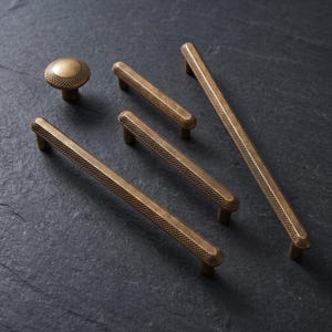 May include: A collection of antique brass hardware including one round knob and four linear drawer pulls of varying lengths. Each piece features a consistent textured knurled pattern across the surface, providing a tactile grip and metallic finish.