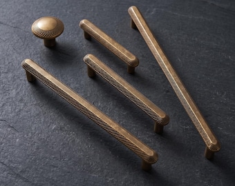 Può includere: Un set di ferramenta in ottone anticato composto da un pomello rotondo e quattro maniglie lineari per cassetti di varie lunghezze. Ogni pezzo presenta un motivo zigrinato testurizzato uniforme sulla superficie, che offre una presa tattile.