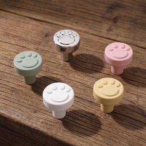 May include: Five round cabinet knobs featuring a raised paw print design on the top surface. The set includes one metallic silver knob, one matte white knob, one sage green knob, one soft pink knob, and one mustard yellow knob.