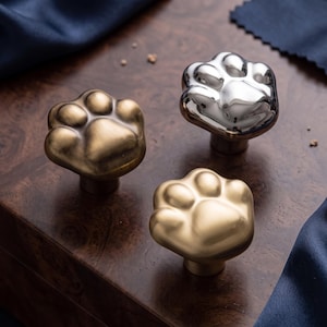 May include: Three cabinet knobs shaped like animal paws, featuring one brushed brass finish, one polished chrome finish, and one antique bronze finish. Each knob has a central pad and four toe pads, designed for attachment to furniture or cabinetry.