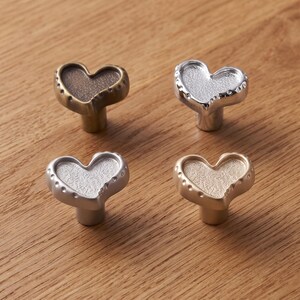 May include: Four heart-shaped cabinet knobs arranged in a square. Each knob features a textured center and a smooth, rounded border with small indentations. The finishes include antique brass, polished silver, matte silver, and champagne gold.