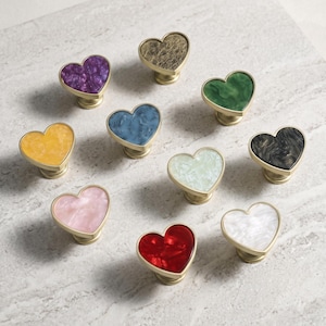 May include: A collection of ten heart-shaped cabinet knobs featuring a gold-toned metal base and a smooth, marbled acrylic top in various colors including purple, gold, green, yellow, blue, black, white, pink, red, and off-white.