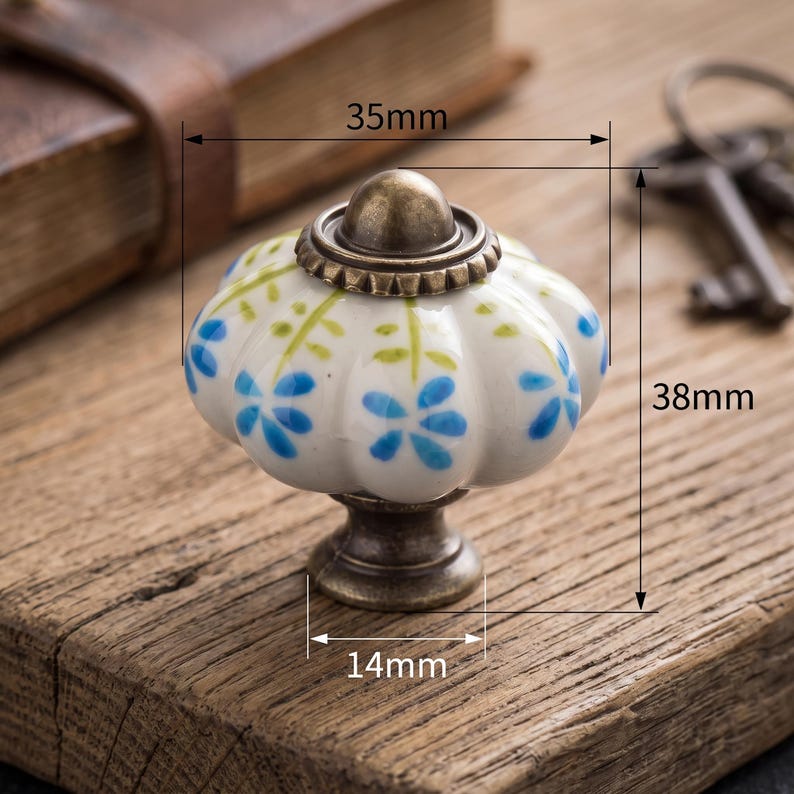 Pu&ograve; includere: Un pomello per mobili in ceramica bianca a forma di zucca scanalata, decorato con fiori blu e steli verdi dipinti a mano. Presenta una base e un tappo in metallo color bronzo antico. Le dimensioni sono 35mm di larghezza, 38mm di altezza e 14mm.