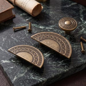 May include: Two semi-circular brass-toned cabinet pulls and one circular brass-toned cabinet knob, each featuring an intricate geometric border pattern and a central textured design, accompanied by several small metal mounting screws.