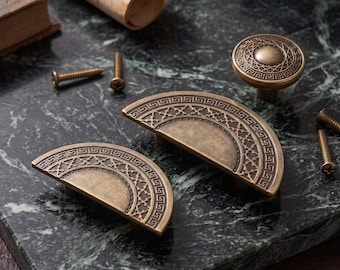 Può includere: Due maniglie per armadietto semicircolari e un pomello circolare in ottone, ciascuno caratterizzato da un intricato motivo geometrico sul bordo e un design centrale testurizzato, accompagnati da diverse piccole viti di montaggio.