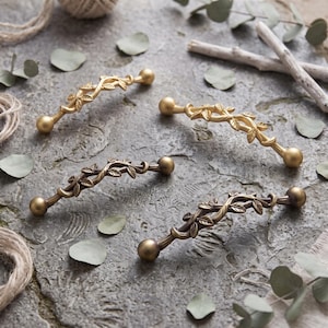 May include: Four metal cabinet pulls arranged in a group. Two pulls feature a bright gold finish, while the other two have a darker, antique bronze finish. Each pull has a central vine and leaf design with spherical ends for mounting.