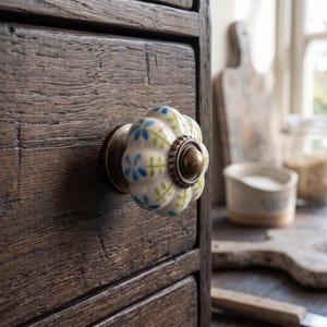 Pu&ograve; includere: Un pomello per cassetto in ceramica rotondo e scanalato, caratterizzato da una base bianca con un motivo dipinto a mano di piccoli fiori blu e steli verdi. Il pomello &egrave; fissato a un frontale di cassetto in legno scuro e usurato con una base in metallo.
