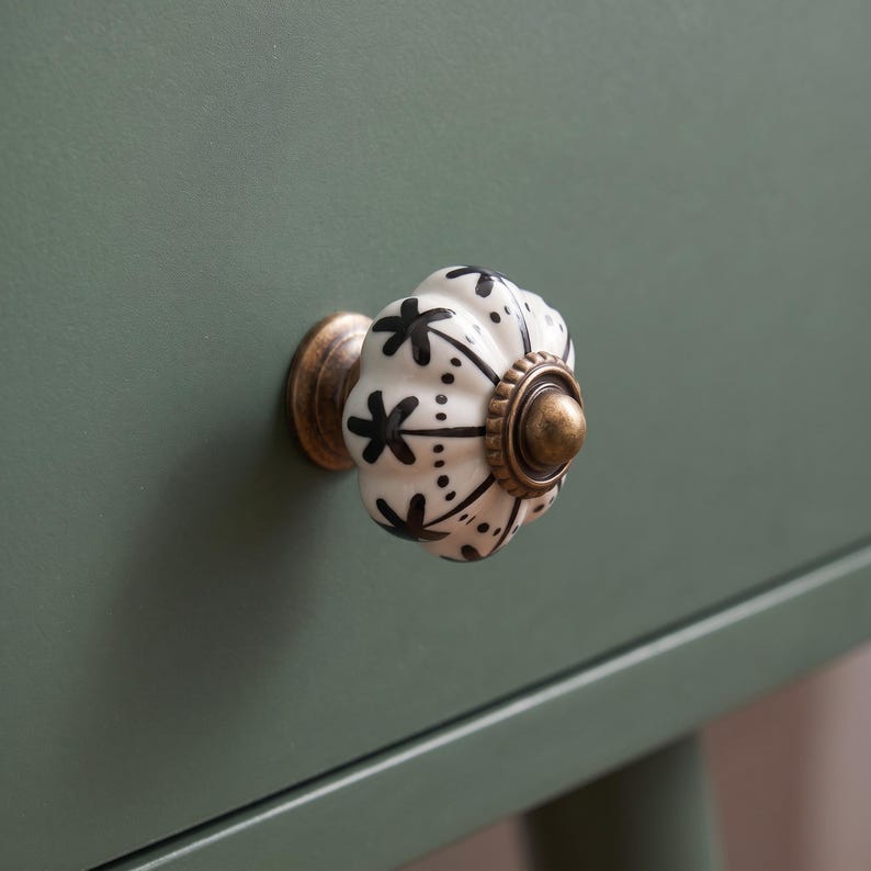 Pu&ograve; includere: Un pomello per cassetto in ceramica rotondo e scanalato, caratterizzato da una base bianca con motivi floreali e puntini dipinti a mano in nero. Il pomello &egrave; montato su una base in metallo color ottone con un accento centrale, su superficie verde oliva.