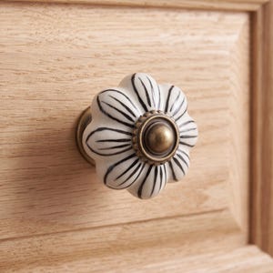 Pu&ograve; includere: Un pomello per mobili a forma di fiore con una superficie in ceramica bianca, dettagli a petalo dipinti in nero e un accento circolare centrale in metallo color ottone antico, montato su un cassetto in legno chiaro con venature visibili.