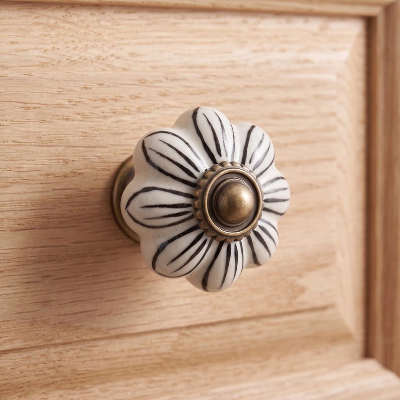 Pu&ograve; includere: Un pomello per mobili a forma di fiore con una superficie in ceramica bianca, dettagli a petalo dipinti in nero e un accento circolare centrale in metallo color ottone antico, montato su un cassetto in legno chiaro con venature visibili.