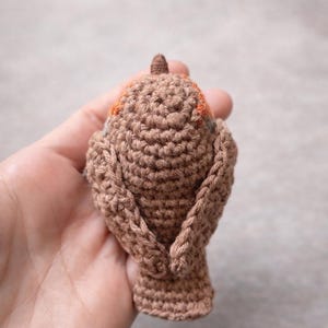 May include: A small, hand-crocheted bird figurine crafted from brown yarn. The bird features a textured, stitched surface with a distinct orange patch visible near the head area, a small pointed beak, and folded wings resting against its body.