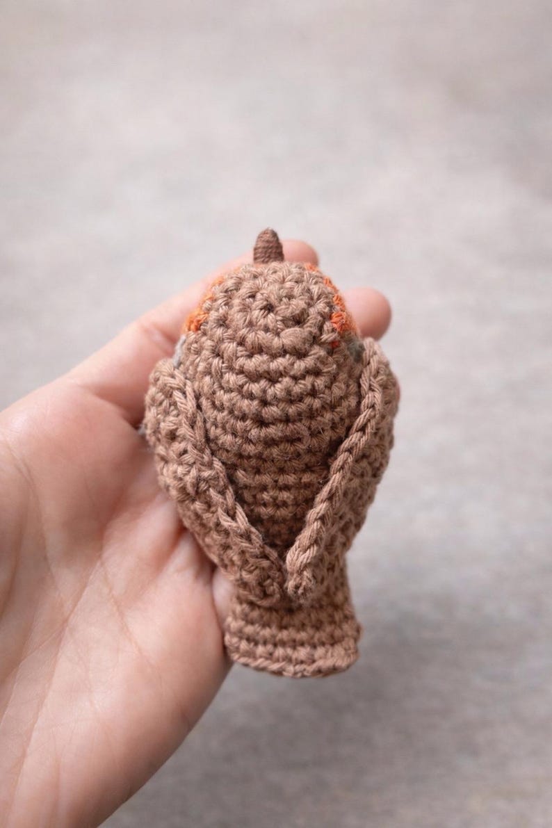May include: A small, hand-crocheted bird figurine crafted from brown yarn. The bird features a textured, stitched surface with a distinct orange patch visible near the head area, a small pointed beak, and folded wings resting against its body.