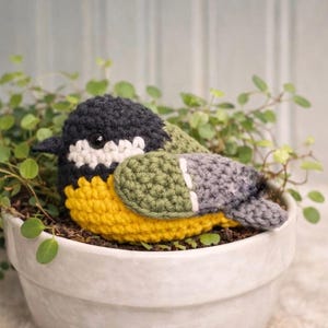 May include: A crocheted bird figurine featuring a black head, white cheek patches, a bright yellow chest, and olive green and grey wings. The bird has a small black beak and a single visible dark eye, crafted with textured yarn stitches.