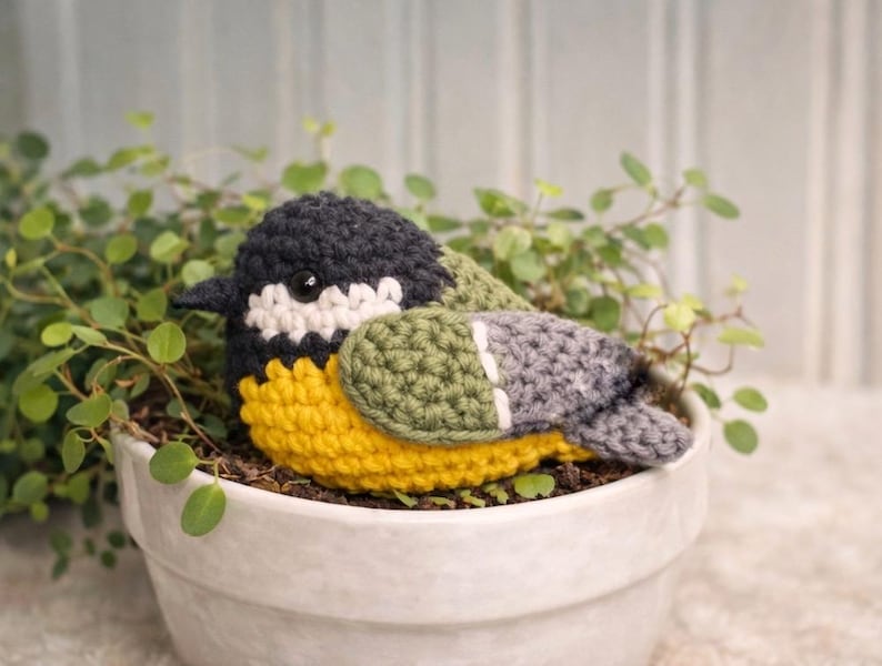 May include: A crocheted bird figurine featuring a black head, white cheek patches, a bright yellow chest, and olive green and grey wings. The bird has a small black beak and a single visible dark eye, crafted with textured yarn stitches.