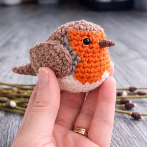 May include: A small, round, crocheted bird figurine featuring a brown back and wings, a bright orange chest, a white belly, and a dark brown beak. The bird has a single visible black bead eye and is crafted with detailed yarn stitching.