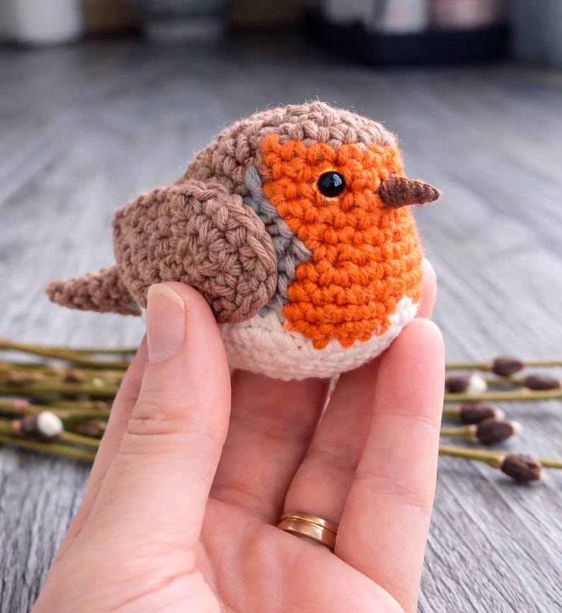 May include: A small, round, crocheted bird figurine featuring a brown back and wings, a bright orange chest, a white belly, and a dark brown beak. The bird has a single visible black bead eye and is crafted with detailed yarn stitching.