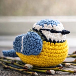 May include: A small, round, crocheted bird figurine featuring a bright yellow belly, blue wings with white accents, and a white face with a black eye and a dark blue stripe across the head. The bird has a small, pointed black beak and a blue tail.