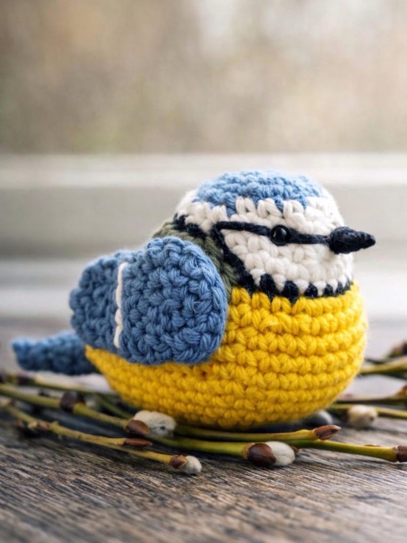 May include: A small, round, crocheted bird figurine featuring a bright yellow belly, blue wings with white accents, and a white face with a black eye and a dark blue stripe across the head. The bird has a small, pointed black beak and a blue tail.