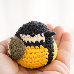 May include: A small, round, crocheted bird figurine featuring a black head, a white cheek patch, a bright yellow belly, and a dark olive green wing. The bird has a small, dark, glossy bead for an eye and a tiny, pointed, dark-colored beak.