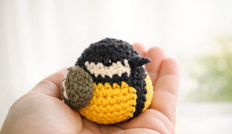 May include: A small, round, crocheted bird figurine featuring a black head, a white cheek patch, a bright yellow belly, and a dark olive green wing. The bird has a small, dark, glossy bead for an eye and a tiny, pointed, dark-colored beak.