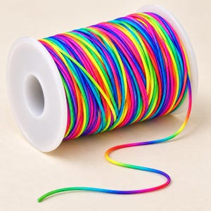 May include: A spool of multi-colored cord featuring a vibrant rainbow gradient pattern of neon pink, yellow, green, and blue. The cord is tightly wound around a white plastic cylindrical spool, with a single strand extending from the main coil.