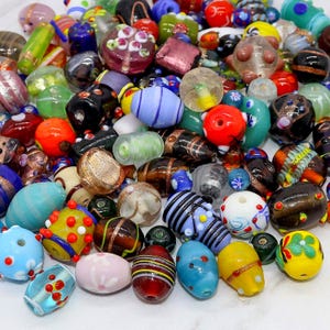 Puede incluir: Una colección de coloridas cuentas de vidrio de varias formas y tamaños. Las cuentas presentan una variedad de colores, incluyendo rojo, azul, amarillo y verde, con patrones y diseños decorativos. Algunas cuentas son redondas, otras ovaladas o cilíndricas.