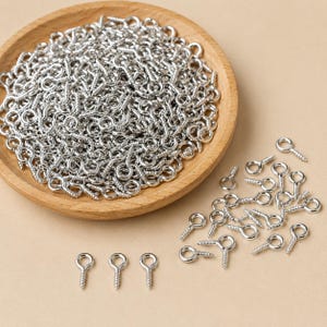 May include: A large collection of silver-toned metal eye screws, each featuring a circular loop at the top and a threaded shank at the bottom. The majority of the eye screws are contained within a round wooden bowl, with several scattered outside.