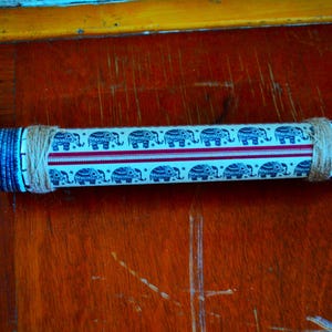 Op de afbeelding: Een cilindervormige caleidoscoop met een witte achtergrond, versierd met blauwe olifantenpatronen en een rode streep. De uiteinden zijn omwikkeld met touw en hebben een blauw patroon. De caleidoscoop ligt op een houten oppervlak.