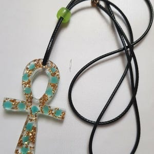 Könnte beinhalten: Ankh-Anhänger-Halskette mit schwarzem Band. Der Ankh ist transparent mit Goldflocken und türkisfarbenen Akzenten. Eine kleine grüne Perle befindet sich auf dem Band. Die Halskette hat einen goldfarbenen Verschluss.