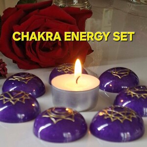 Może przedstawiać: Zestaw Chakra Energy z siedmioma fioletowymi kamieniami ze złotymi wzorami, ułożonymi wokół zapalonej świecy typu tealight. Czerwona róża i inne elementy kwiatowe w tle. Wyświetlany jest tekst "CHAKRA ENERGY SET".