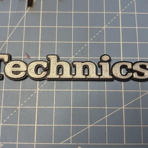 Puede incluir: Logotipo de Technics plateado y negro, con efecto tridimensional en relieve. La palabra "Technics" está en negrita, sin serifa. El logotipo está sobre un fondo de cuadrícula azul.