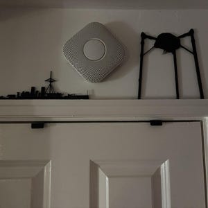 May include: A white and grey smoke detector is mounted on a white wall. A black metal silhouette of a ship and a black metal decorative object are also mounted on the wall above a white door.